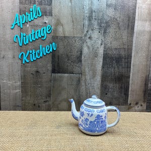 May include: A white and blue ceramic teapot with a lid, featuring a scenic design. The teapot has a curved spout and handle. The text "Aprils Vintage Kitchen" is above the teapot. The teapot is on a tan surface with a wooden background.