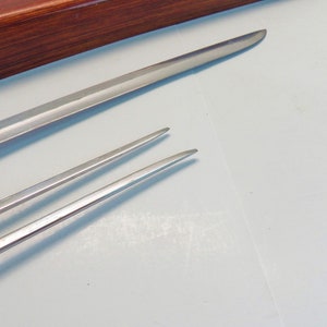 May include: Close-up of two stainless steel serving utensils. One is a long, slender knife, and the other is a two-pronged fork. The utensils are on a white surface, with a wooden box in the background.