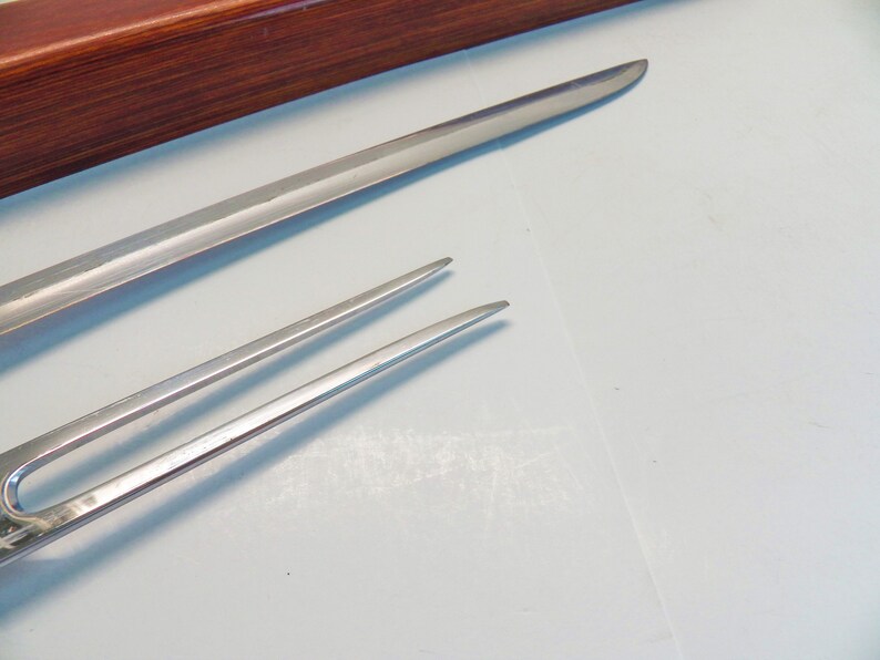May include: Close-up of two stainless steel serving utensils. One is a long, slender knife, and the other is a two-pronged fork. The utensils are on a white surface, with a wooden box in the background.