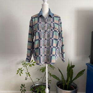 May include: A long-sleeved button-up shirt with a patchwork design in shades of blue, green, pink, and beige. The shirt has a denim collar and two flap pockets.