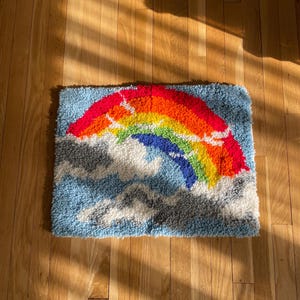 May include: A rectangular rug featuring a colorful rainbow design against a light blue background with white and gray cloud accents. The rainbow displays vibrant hues of red, orange, yellow, green, and blue.