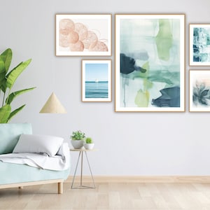 Op de afbeelding: Een verzameling ingelijste kunstdrukken tentoongesteld op een witte muur. De prints tonen abstracte aquarelontwerpen in blauwe, groene en perzikkleuren, samen met een zeegezicht en botanische kunst. De lijsten zijn licht van kleur.