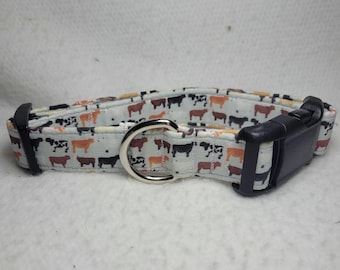 May include: A dog collar with a repeating pattern of black, brown, and orange cows on a white background. The collar has a black plastic buckle and a silver metal D-ring.
