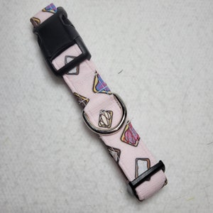 May include: A light pink dog collar with a repeating pattern of colorful pastries. The collar has a black plastic buckle and a silver metal D-ring. The collar is designed for a small to medium-sized dog.
