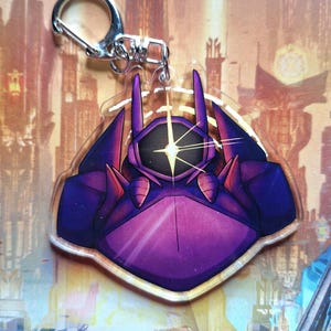 May include: A purple and black robot-themed keychain with a silver clasp. The keychain features a stylized robot design with a starburst detail. The robot has a purple body with black and red accents. The background shows a futuristic city.