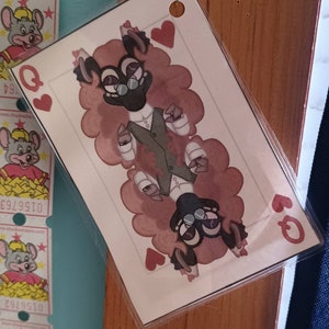 May include: A playing card featuring a cartoon character with a brown furry body, a black face, and glasses. The character is wearing a green vest and white shirt. The card is a Queen of Hearts.