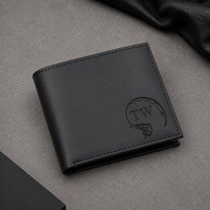 K&ouml;nnte beinhalten: Ein schwarzes Klappportemonnaie mit einer gepr&auml;gten kreisf&ouml;rmigen Grafik in der vorderen Ecke. Das Design zeigt die Buchstaben TW innerhalb eines kreisf&ouml;rmigen Angelmotivs mit einem Angelhaken und einem Fisch. Sichtbare N&auml;hte.