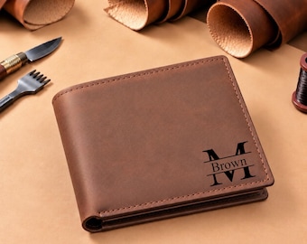 Puede incluir: Una cartera bifold de cuero marrón con un diseño de monograma grabado oscuro que presenta la letra M en la parte superior e inferior, y el nombre Brown centrado horizontalmente entre las letras. La cartera tiene costuras visibles en los bordes.