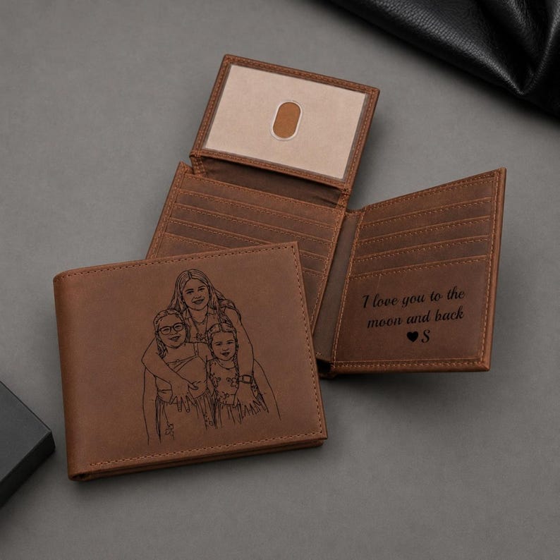 K&ouml;nnte beinhalten: Ein braunes Leder-Bifold-Portemonnaie mit einer lasergravierten Strichzeichnung von drei Personen auf der Vorderseite. Das Innere enth&auml;lt Kartenf&auml;cher und ein Sichtfenster. Auf der rechten Seite ist der Text I love you to the moon and back S eingraviert.