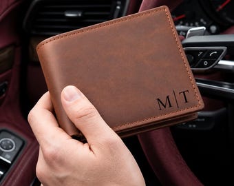 Puede incluir: Una cartera plegable de color marrón con un acabado liso y mate y costuras visibles en los bordes. La esquina inferior derecha presenta las letras grabadas M y T separadas por una línea vertical, creando un diseño de monograma.