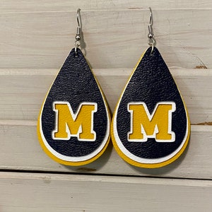 Op de afbeelding: Een paar traanvormige leren oorbellen met een geel, wit en donkerblauw ontwerp. De oorbellen hebben een grote, gele "M" in een witte omtrek.