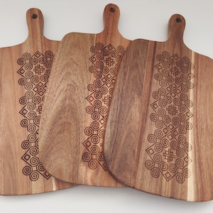 May include: Three wooden cutting boards with a decorative geometric pattern engraved on the surface. The boards have a handle on the top and are made of light brown wood.
