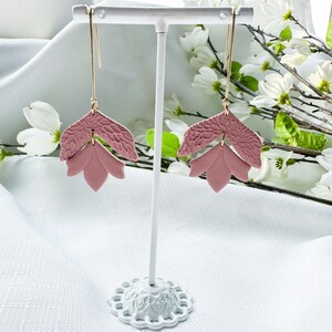 May include: A pair of pink floral dangle earrings with gold hooks. The earrings are displayed on a white stand.