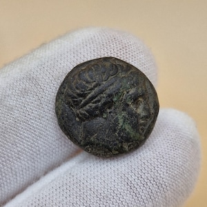 May include: A small, round, antique bronze coin. The coin shows a detailed profile of a person with a laurel wreath. The coin has a dark, aged patina with hints of green oxidation.