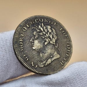 Può includere: Una moneta di bronzo con l'iscrizione "IN MEMORY OF GEORGE THE FOURTH" attorno al bordo e un profilo di un uomo con una corona di alloro sulla testa.