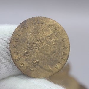 May include: A bronze-colored coin featuring a profile of a person with a laurel wreath and text around the edge. The coin has visible wear and tear, suggesting age. The coin's design includes the inscription "GEORGIUS II DEI GRATIA."