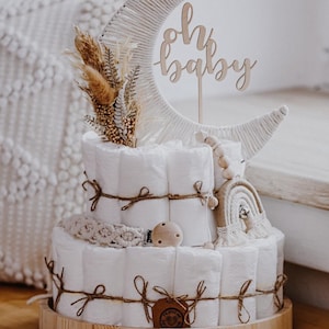 Könnte beinhalten: Eine zweistöckige Windeltorte, verziert mit einem Holzmond und den Worten "oh baby". Die Torte ist mit Schnur umwickelt und mit Holzperlen, einem Makramee-Regenbogen und einem Holz-Beißring verziert.