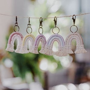 May include: Five rainbow-colored macrame keychains with a white tassel. The keychains are hanging from a string.