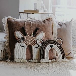 Op de afbeelding: Twee macramé wandhangers in de vorm van regenbogen. Eén is bruin en wit met een hertengezicht, de andere is bruin en wit met een berengezicht. De hangers worden getoond op een bruine bank met een bruin kussen.