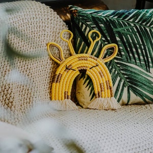 Puede incluir: Un arcoíris de macramé amarillo y marrón con un diseño de cabeza de jirafa. El arcoíris está colgado de una manta de punto blanco.