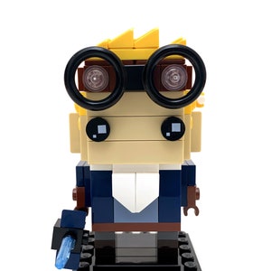 May include: A Lego minifigure of a person with blond hair, wearing round black glasses, a blue jacket, and brown pants. The minifigure is holding a blue lightsaber in its right hand and is standing on a black base.