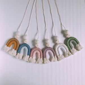 May include: A collection of handmade macrame rainbow wall hangings in various colors, including mustard yellow, blue, pink, and green. Each rainbow has a white fringe and is suspended by a natural cord with a wooden bead and a felt ball.