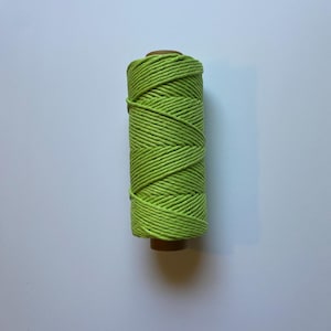 May include: A spool of bright green macrame cord. The cord is tightly wound around a brown cardboard core. The color is a vibrant, spring-like green, and the texture of the cord is visible.