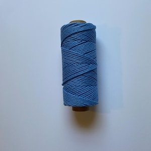 May include: A spool of blue cotton cord. The cord is tightly wound around a cardboard core. The color is a medium blue, and the texture of the cord is visible. The spool is sitting on a white surface.