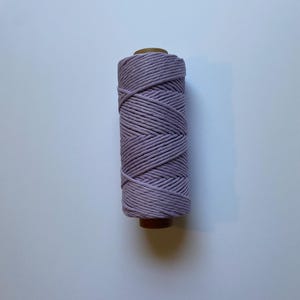 May include: A spool of lavender-colored macrame cord. The cord is tightly wound around a brown cardboard core. The texture of the cord is visible, with each strand neatly aligned. The background is a plain white surface.