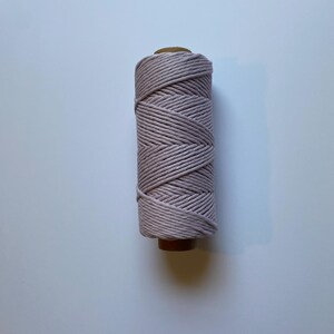 May include: A spool of light pink macrame cord. The cord is tightly wound around a brown cardboard core. The color is a soft, muted pink, and the texture of the cord appears to be smooth and slightly twisted. The spool is set against a white background.