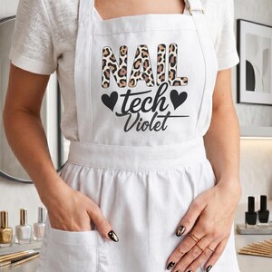 May include: A white apron featuring a ruffled hem and two front pockets. The chest area displays the text "NAIL" in a leopard print pattern, followed by the black text "tech" and "Violet" with two black heart icons on either side.