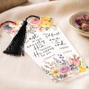 May include: A clear rectangular bookmark featuring a floral design with pink, yellow, and purple flowers. The text reads: Cast all your anxiety on Him because He cares for you - 1 Peter 5:7 and The steadfast love of the Lord never ceases ns 3:22-23. It has a black tassel.