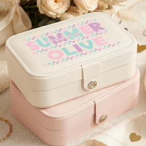 May include: Two stacked jewelry boxes, one cream and one pink, with silver clasps. The top box has the words "SUMMER OLIVE" in pastel colors. A gold bracelet and pearl earrings are visible in the foreground, suggesting a jewelry theme.