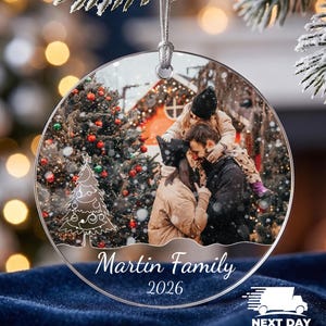 May include: A round, clear acrylic Christmas ornament featuring a printed photograph of a family embracing outdoors in the snow. The ornament includes a white line-art Christmas tree, the text Martin Family, and the year 2026 in white script.