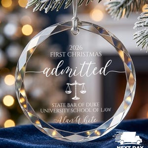 May include: A circular clear glass ornament with faceted edges, featuring white engraved text that reads 2026 FIRST CHRISTMAS admitted, a scale of justice icon, STATE BAR OF DUKE UNIVERSITY SCHOOL OF LAW, and the name Alan White.