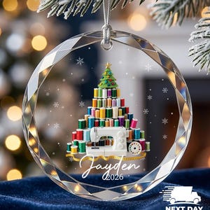 May include: A round, faceted clear glass ornament featuring a printed graphic of a Christmas tree constructed from colorful sewing thread spools, a white sewing machine, and a yellow measuring tape. The name Jayden and the year 2026 are printed below.