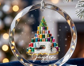 May include: A round, faceted clear glass ornament featuring a printed graphic of a Christmas tree constructed from colorful sewing thread spools, a white sewing machine, and a yellow measuring tape. The name Jayden and the year 2026 are printed below.