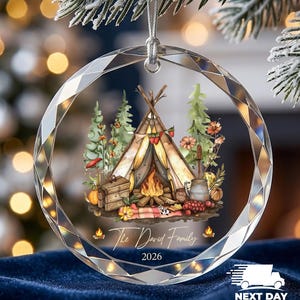 May include: A circular faceted glass ornament featuring a printed illustration of a teepee, a campfire, pine trees, and autumn foliage. The text "The David Family" and "2026" is printed in white script and sans-serif font below the illustration.