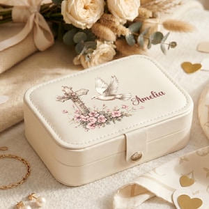 May include: A rectangular cream-colored jewelry box with a snap closure. The lid features a printed design of a cross, a flying white dove, pink flowers, and the name Amelia in script. The box is made of a smooth material with visible stitching.