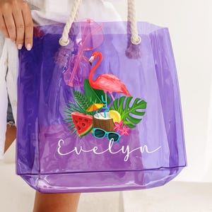 May include: A clear purple tote bag with white rope handles. The bag features a tropical design with a pink flamingo, watermelon, and the name "Evelyn" in white script. Sunglasses are attached to the bag.