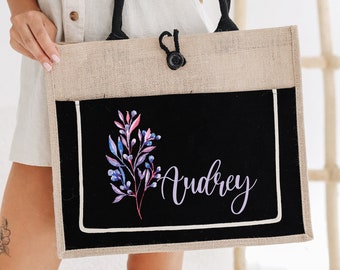 May include: A natural jute tote bag with black handles and a black fabric insert. The insert features a watercolor floral design and the name "Audrey" in purple script.
