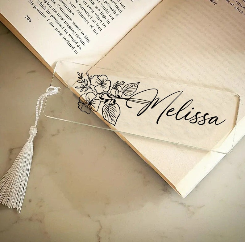 May include: A clear glass bookmark with a black floral design and the name "Melissa" printed in cursive. The bookmark has a white tassel.