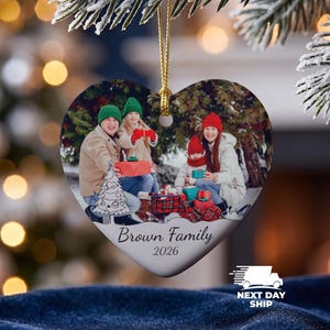 May include: A heart-shaped ceramic Christmas ornament featuring a printed photograph of a family of four outdoors with gifts. The ornament includes a gold hanging cord and the printed text Brown Family 2026 below the photo, with a line drawing of a tree.