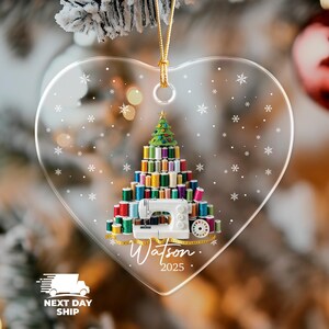 May include: A heart-shaped clear acrylic ornament featuring a graphic of a Christmas tree constructed from colorful sewing thread spools and a white sewing machine. The design includes white snowflakes, a yellow measuring tape, and the text Watson 2025.