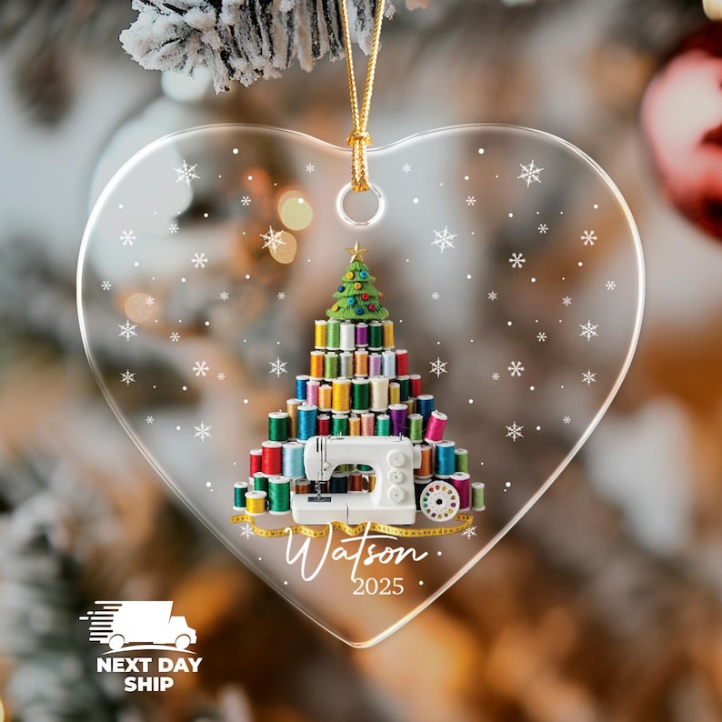 May include: A heart-shaped clear acrylic ornament featuring a graphic of a Christmas tree constructed from colorful sewing thread spools and a white sewing machine. The design includes white snowflakes, a yellow measuring tape, and the text Watson 2025.