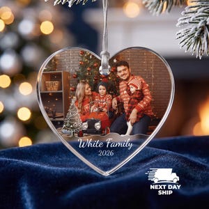 May include: A heart-shaped clear acrylic Christmas ornament featuring a printed photograph of a family of four in red patterned sweaters. The ornament includes a silver hanging ribbon, a white graphic of a Christmas tree, and the text White Family 2026.