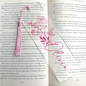 May include: A clear acrylic bookmark with a pink tassel and a floral design. The bookmark is placed on a page of a book with text about crowns and jewels.