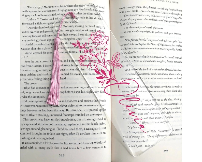 May include: A clear acrylic bookmark with a pink tassel and a floral design. The bookmark is placed on a page of a book with text about crowns and jewels.