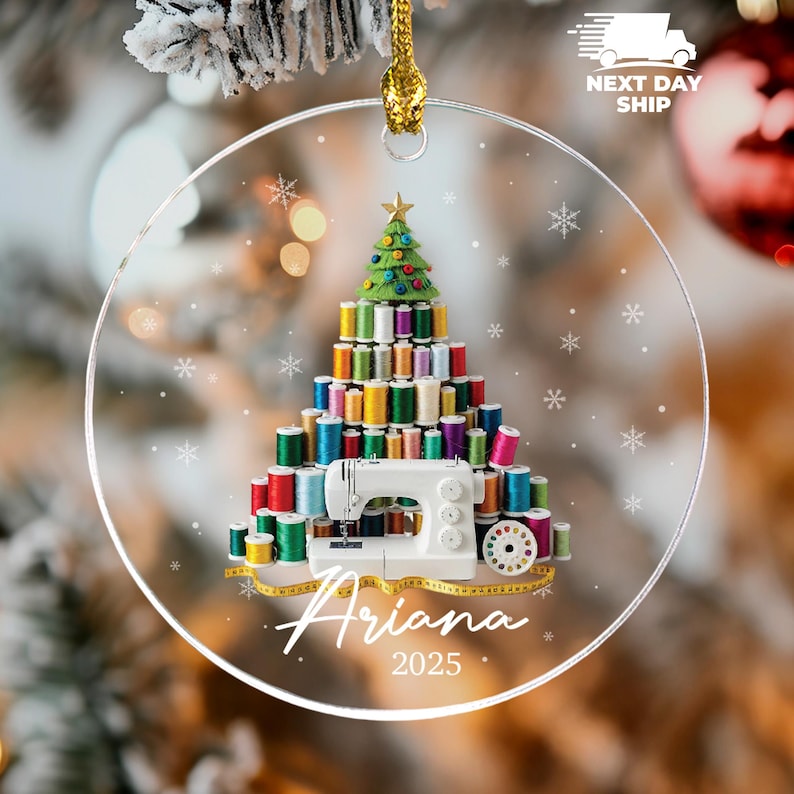 May include: A round, clear acrylic Christmas ornament featuring a printed design of a sewing machine and a tree constructed from colorful thread spools. The ornament includes a yellow measuring tape, the name Ariana, and the year 2025 in white text.