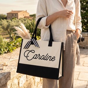 K&ouml;nnte beinhalten: Eine rechteckige Tragetasche aus Segeltuch mit einem cremefarbenen oberen Bereich und einem schwarzen Boden. Der Name Caroline ist in schwarzer Schreibschrift auf der Vorderseite aufgedruckt. Die Tasche hat schwarze Tragegriffe und eine schwarz-wei&szlig; gestreifte Schleife.